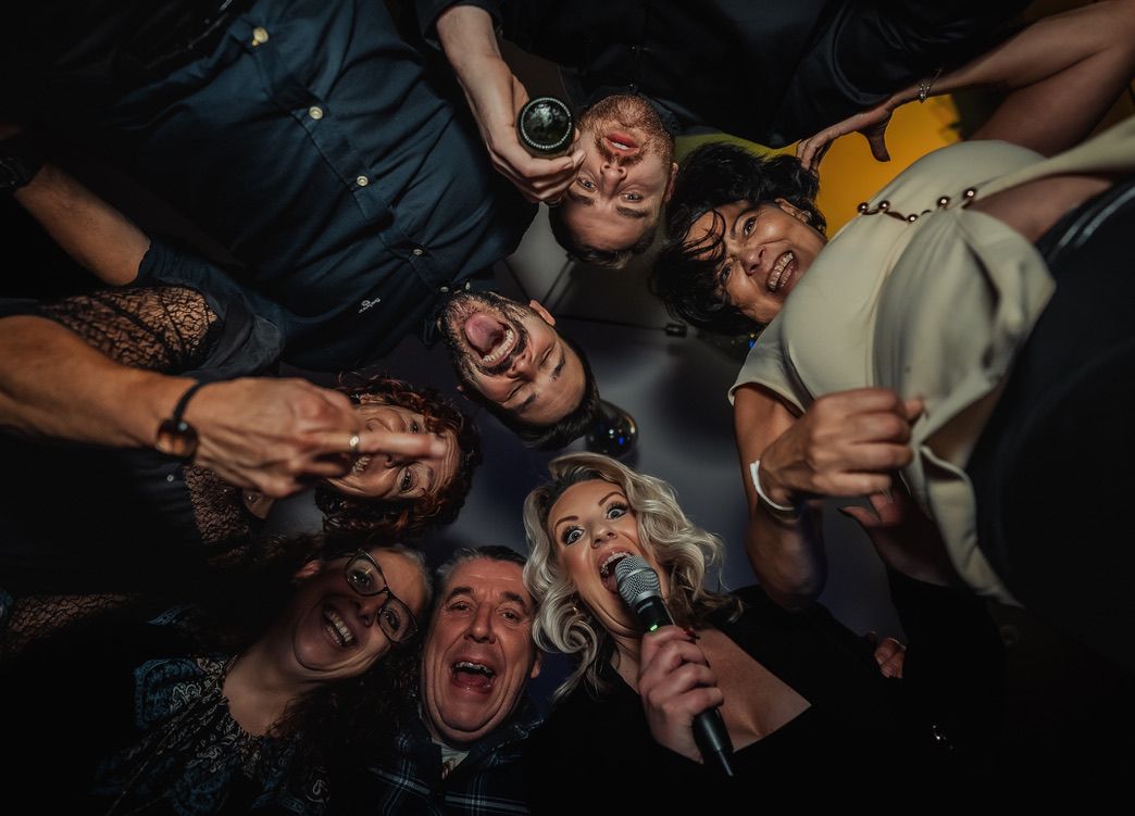 Guests gather around the camera, laughing and singing with a microphone at a party.