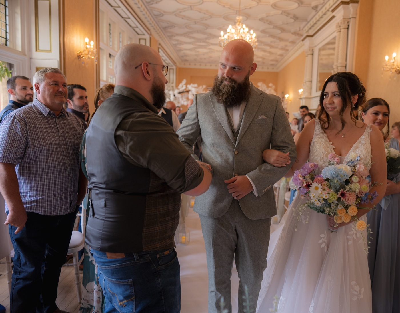 A bride holding a colourful bouquet stands beside a man in a grey suit greeting a guest during the ceremony.