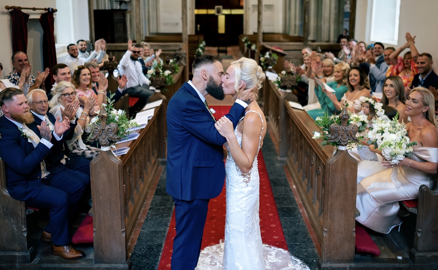 A newly married couple kiss in a church aisle as guests applaud.