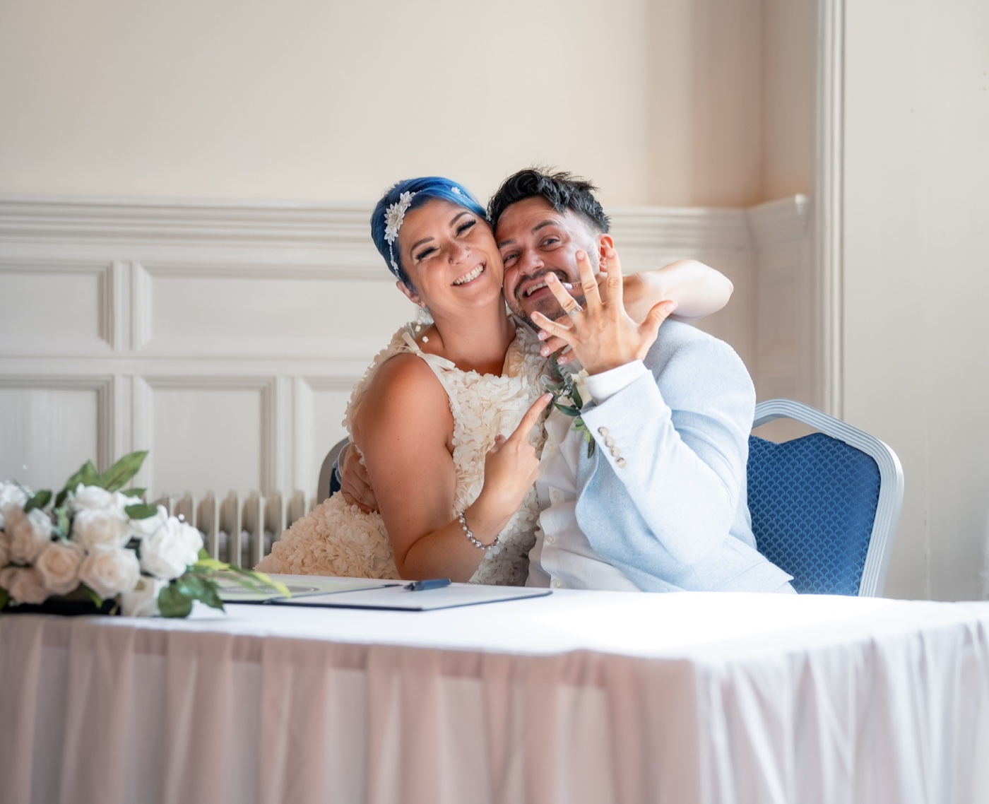A smiling newly married couple show their wedding rings while seated at a signing table.